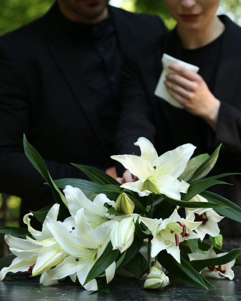 couple-near-granite-tombstone-with-white-lilies-cemetery-outdoors-selective-focus-funeral-ceremony